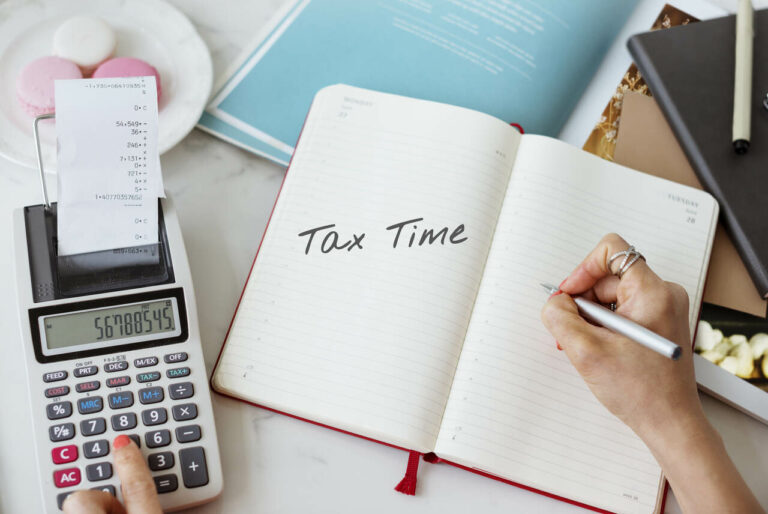 top view of a person writing the word 'tax time' on a note pad with one hand and using calculator in another